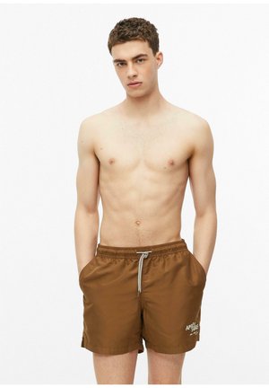 Jeune homme aux cheveux courts et bruns portant un short de bain marron avec poches et cordon, debout devant un fond blanc uni.