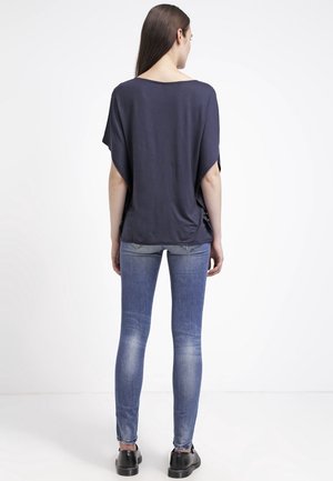 Mujer de espaldas, con una blusa suelta azul marino oscuro, vaqueros ajustados azules desgastados y zapatos negros sobre un fondo blanco liso.