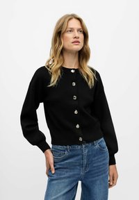 Cardigan nero a maglia con maniche lunghe, caratterizzato da sei bottoni argentati sul fronte. Abbinato a jeans blu per creare contrasto. Texture liscia.