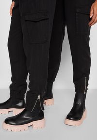 Pantalons cargo noirs avec poches latérales et finitions zippées, associés à des bottes noires chunky avec une semelle plateforme rose. Texture lisse et matériau durable.