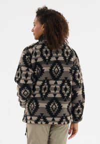 Pull en polaire beige avec des motifs géométriques noirs. Il présente une coupe décontractée, des manches longues et un ourlet arrondi. Texture douce.