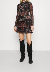 Mustat kuviolliset mekko pitkillä hihoilla, kerroksellinen muotoilu, kukkakuosi punaisena ja vihreänä, yhdistettynä mustiin polvenkorkuisiin löysiin saapikkaisiin.