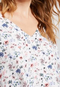Blusa floral con escote en V de tela blanca. Presenta pequeños diseños coloridos de flores rosas, azules y verdes. Textura suave.