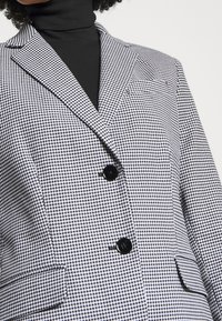 Černobílé sako s vzorem houndstooth, které má klopený límec, dvě černé knoflíky a boční kapsy, vyrobené z texturované tkaniny.