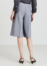 Personne portant un pantalon large gris clair arrivant aux genoux, des sandales à talons noires et une chemise à rayures foncées à manches longues, se tenant sur un sol blanc.