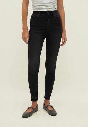 Woman wearing high-waist black skinny jeans with frayed hems and gray Mary Jane flats, standing against a plain light background.