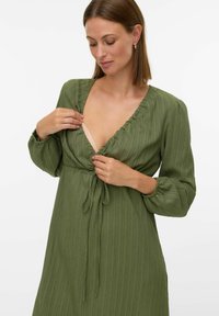 Sieviete pielāgo garās piedurknes zaļās teksturētās kleitas kakla izgriezumu ar sasietu jostu, skatoties uz leju uz viendabīga balta fona.