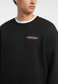 Svart hettegenser laget av bomullsblanding, med rund hals og oversized passform. Brodert "GUESS JEANS" logo i rødt på brystet.