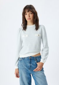 Lichtblauwe gestreepte trui met pofmouwen, voorzien van witte zakken en decoratieve knopen, gecombineerd met blauwe jeans op een effen achtergrond.