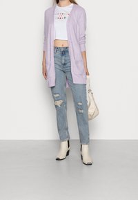Cárdigan abierto de color púrpura claro sobre una camiseta corta blanca con texto colorido, combinado con jeans azules desgastados y botines de tacón crema.