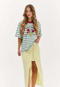 Mladá žena s dlouhými vlnitými vlasy nosí pruhovanou košili s květinovým motivem a žlutou sukni s vysokým rozparkem, na které jsou barevné knoflíky.