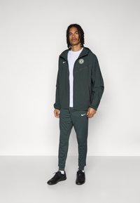 Grønn atletisk jakke med hette, glidelås foran og Nike-logo; parret med matchende bukser og svarte treningssko. Glatt, lett stoff.