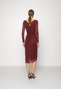 Burgundská midi šaty s dlouhými průhlednými rukávy, nabíraným detailem na zadní části a s vlnkovaným lemem, doplněné černými sandály na vysokém podpatku.