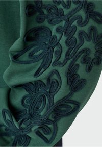 Texto textil verde con patrones florales bordados en verde oscuro, con bucles intrincados y remolinos a lo largo del borde de la tela. Textura suave.