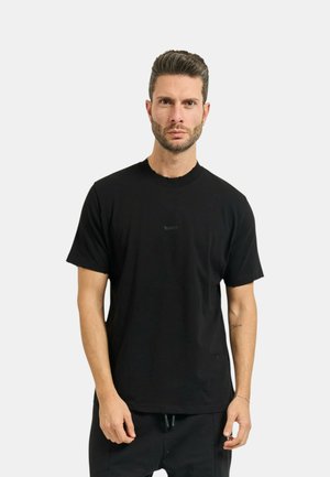 Uomo con capelli corti e barba, che indossa una maglietta nera a maniche corte e pantaloni neri, in piedi davanti a uno sfondo chiaro e uniforme.