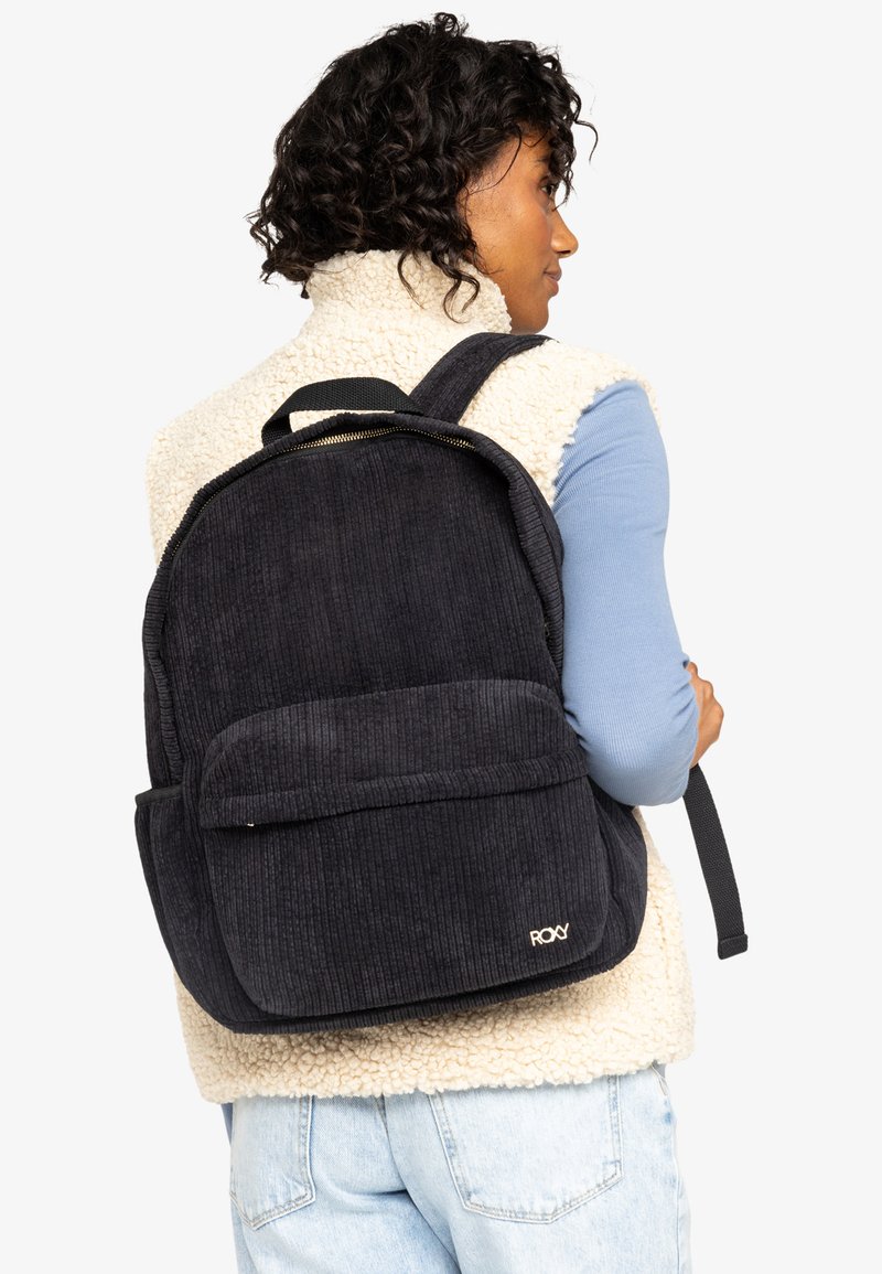 Sac en velours côtelé noir avec une poche avant et des bretelles ajustables, présenté sur un fond blanc avec un contraste de texture.