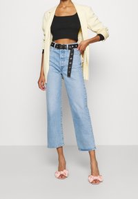 Henkilö, joka on pukeutunut vaaleansinsisiin korkeavyötäröisiin farkkuihin, joissa on musta vyö, mustaan lyhythihaiseen toppiin, kermaiseen bleiseriin ja vaaleanpunaisiin aukileikkureihin, seisoo valkoista seinää vasten.