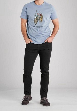 Uomo che indossa una t-shirt azzurra chiara della U.S. Polo Assn. con una grafica di giocatori di polo, pantaloni neri slim e scarpe stringate marroni scure, in piedi con le mani in tasca.