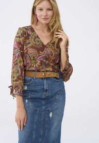Bruine blouse met een patroon en groene en roze details, V-hals en uitlopende mouwen, gecombineerd met een spijkerrok met rafelaccenten en een bruine riem.
