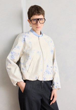 Osoba s krátkymi vlasmi a veľkými okuliarmi, ktorá má na sebe svetlú kvetinovú bundu na zips cez modrú košeľu a tmavé nohavice, stojaca pri obyčajnej stene.