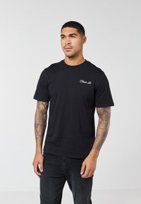 Άντρας με κοντά μαλλιά και τατουάζ σε όλο το μήκος του βραχ�ίονα φοράει μαύρο μπλουζάκι με σχέδιο και μαύρο παντελόνι, στέκεται μπροστά σε απλό ανοιχτό φόντο.