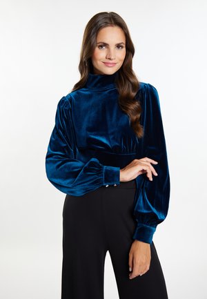 Samtoberteil in tiefblau mit hohem Kragen, Puffärmeln und gefütterten Handgelenken. Glatte Textur, kombiniert mit schwarzen hochtaillierten Hosen.