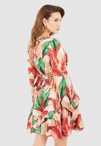 Abito floreale con sfondo beige, caratterizzato da grandi motivi di foglie rosse e verdi, maniche lunghe, vita cintrata e orlo a strati.