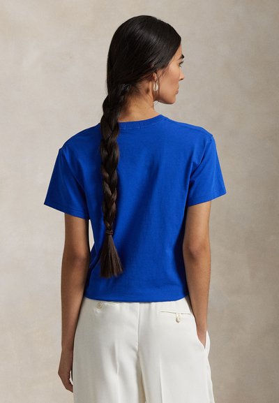 Sieviete ar gariem pītiem matiem valkā zilu īss piedurkņu t-kreklu un baltas bikses, stāvot ar muguru pret kameru.