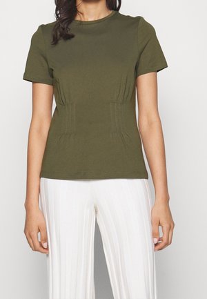 Frau trägt ein olivgrünes, kurzärmeliges, tailliertes T-Shirt mit vertikalen Nahtdetails, kombiniert mit weißen, gerippten, weit geschnittenen Hosen.