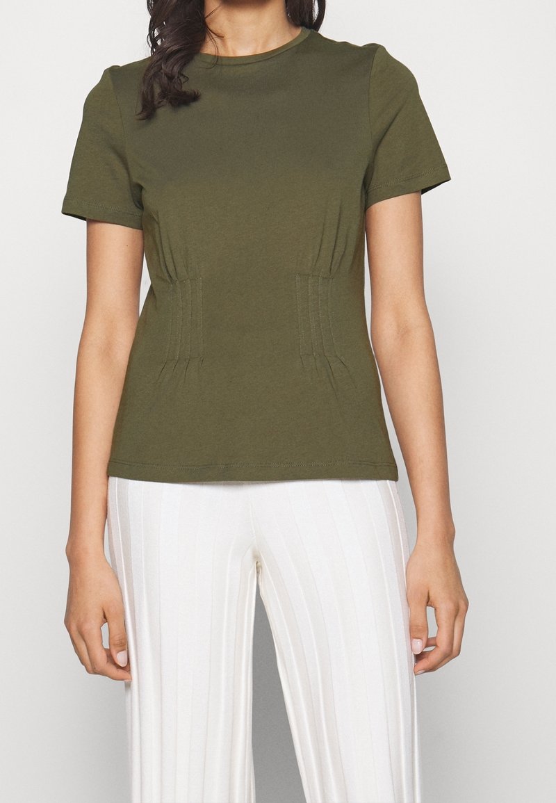 Femme portant un t-shirt ajusté manches courtes vert olive avec des détails de coutures verticales, associé à un pantalon blanc large côtelé.