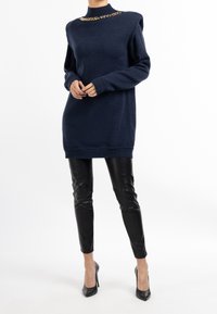 Tmavě modré svetrové šaty s vysokým límcem a zlatým řetízkovým detailem na límci. Kombinované s lesklými černými koženými kalhotami a podpatky.