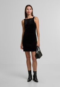 Černé sametové šaty bez rukávů se čtvercovým výstřihem, přiléhavého střihu a hladké textury. Modelka drží malou černou kabelku.