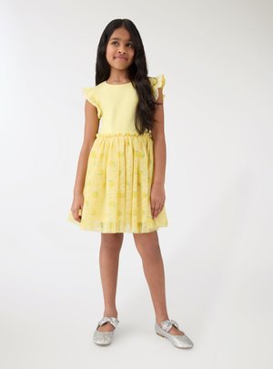 Ragazza con capelli lunghi e scuri che indossa un vestito giallo con maniche arricciate e scarpe argentate glitterate, in piedi su uno sfondo bianco semplice.