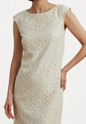 Beżowa koronkowa sukienka bez rękawów z kwiatowymi wzorami, noszona przez osobę z jedną ręką na biodrze, a drugą swobodnie opuszczoną wzdłuż ciała.