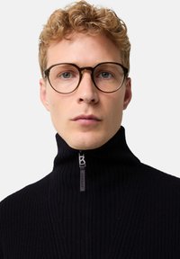 Junger Mann mit lockigem blondem Haar, der runde Brillen und einen schwarzen, gerippten Rollkragenpullover mit Reißverschluss am Kragen trägt.