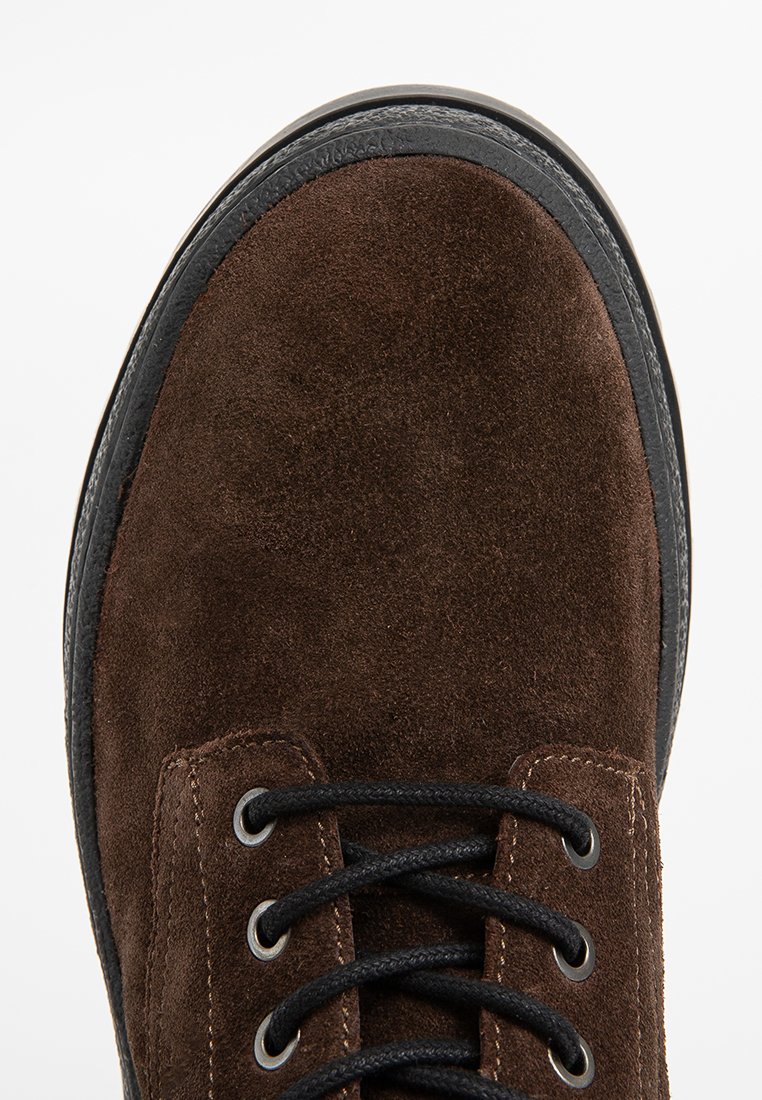 Punta de zapato de cuero de ante marrón con cordones negros y suela de goma negra, visto desde arriba sobre un fondo blanco.