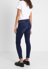 Jeans ajustados de un azul oscuro, con un diseño de cinco bolsillos y detalles de costura sutiles, combinados con una camiseta blanca básica y zapatos negros.
