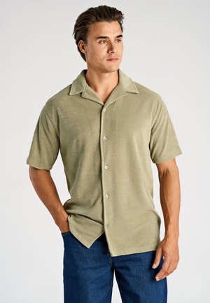 Man met een lichtgroene korte mouwen overhemd met kraag en donkeblauwe spijkerbroek, staand met één hand in de zak, kijkend naar rechts.