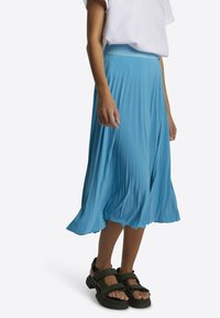 Falda midi plisada de color azul cielo con cinturilla tejida. Combinada con sandalias negras de tiras con suela gruesa.
