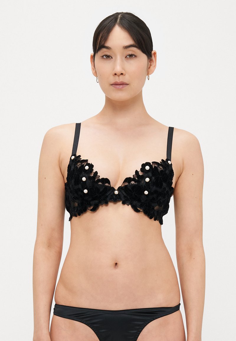 Črni čipkasti modrček z okrasnimi cvetličnimi aplikacijami, dekorativnimi biseri, globokim izrezom in tankimi nastavljivimi naramnicami. Gladke črne spodnjice dopolnjujejo videz.