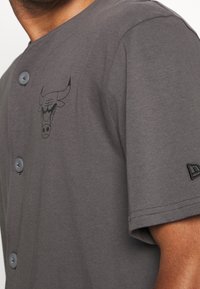 Šedá košile s krátkými rukávy a zapínáním na knoflíky, s logem býka a vyšívaným detailem na rukávu. Měkký bavlněný materiál.