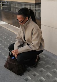 Beige, high-collared jacket with a zipper; black pants; brown suede bag; person crouching on a textured stone surface. Sunlight accentuates the scene.