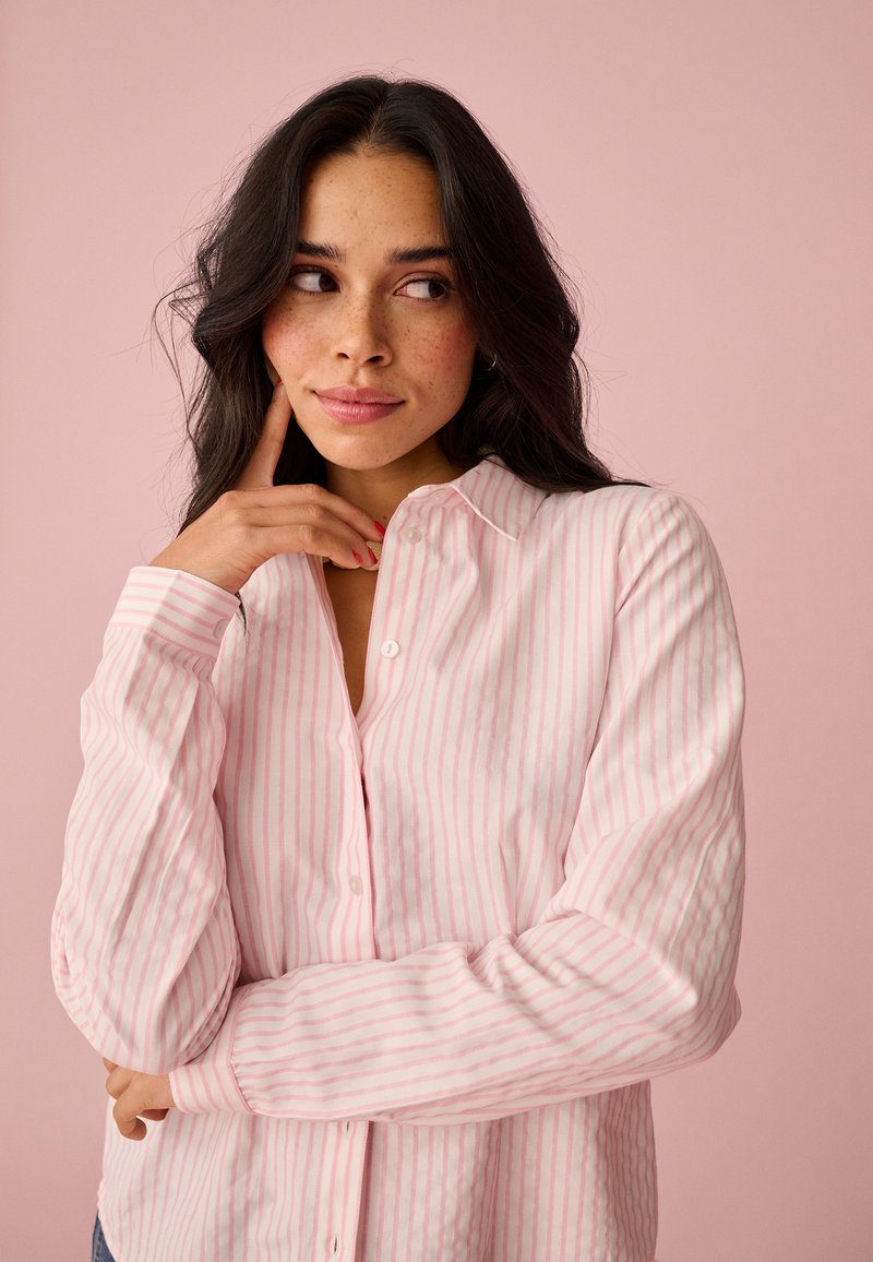 Junge Frau mit langen, dunklen Haaren, die ein rosa-weiß gestreiftes Hemd trägt und nachdenklich vor einem hellrosa Hintergrund posiert.