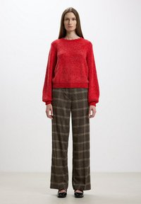 Maglione rosso lavorato a maglia con maniche lunghe, abbinato a pantaloni a gamba larga in tessuto scozzese nei toni del marrone. Scarpe nere flat completano l'outfit.