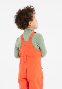 Orange vattentäta overaller med justerbara remmar, i kombination med en grön fleece-tröja. Outfiten har en bakre midjeresår för ökad komfort.