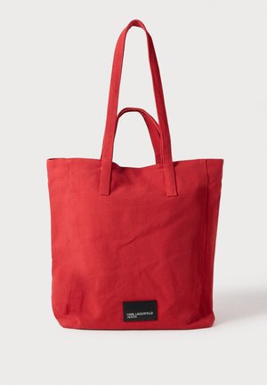 Saco tote de lona vermelho com duas alças de ombro, formato quadrado e uma etiqueta preta na parte inferior com texto branco. Tecido liso, sem textura.