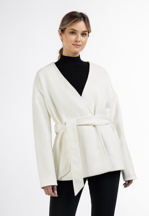 Junge Frau trägt eine weiße, mit Gürtel versehene Wickeljacke über einem schwarzen Rollkragenpullover und schwarzen Hosen, stehend vor einem schlichten weißen Hintergrund.