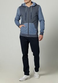 Sudadera con capucha y cremallera en tonos azules, con un gran bolsillo frontal, texturas contrastantes y cordones en la capucha. Combinada con pantalones oscuros.
