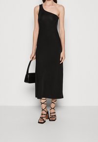 Kobieta ubrana w czarną midi sukienkę na jedno ramię, w czarnych sandałach na wysokim obcasie i trzymająca czarną torebkę na jednolitym tle.