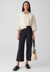 Chemise crème surdimensionnée avec deux poches avant, associée à un pantalon large noir. Les accessoires comprennent un petit sac en osier beige et des mocassins noirs.